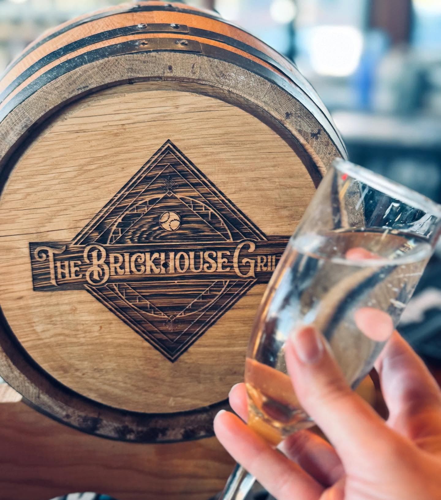 Hand holding a glass of water before a wooden barrel featuring The Brickhouse Grill logo.