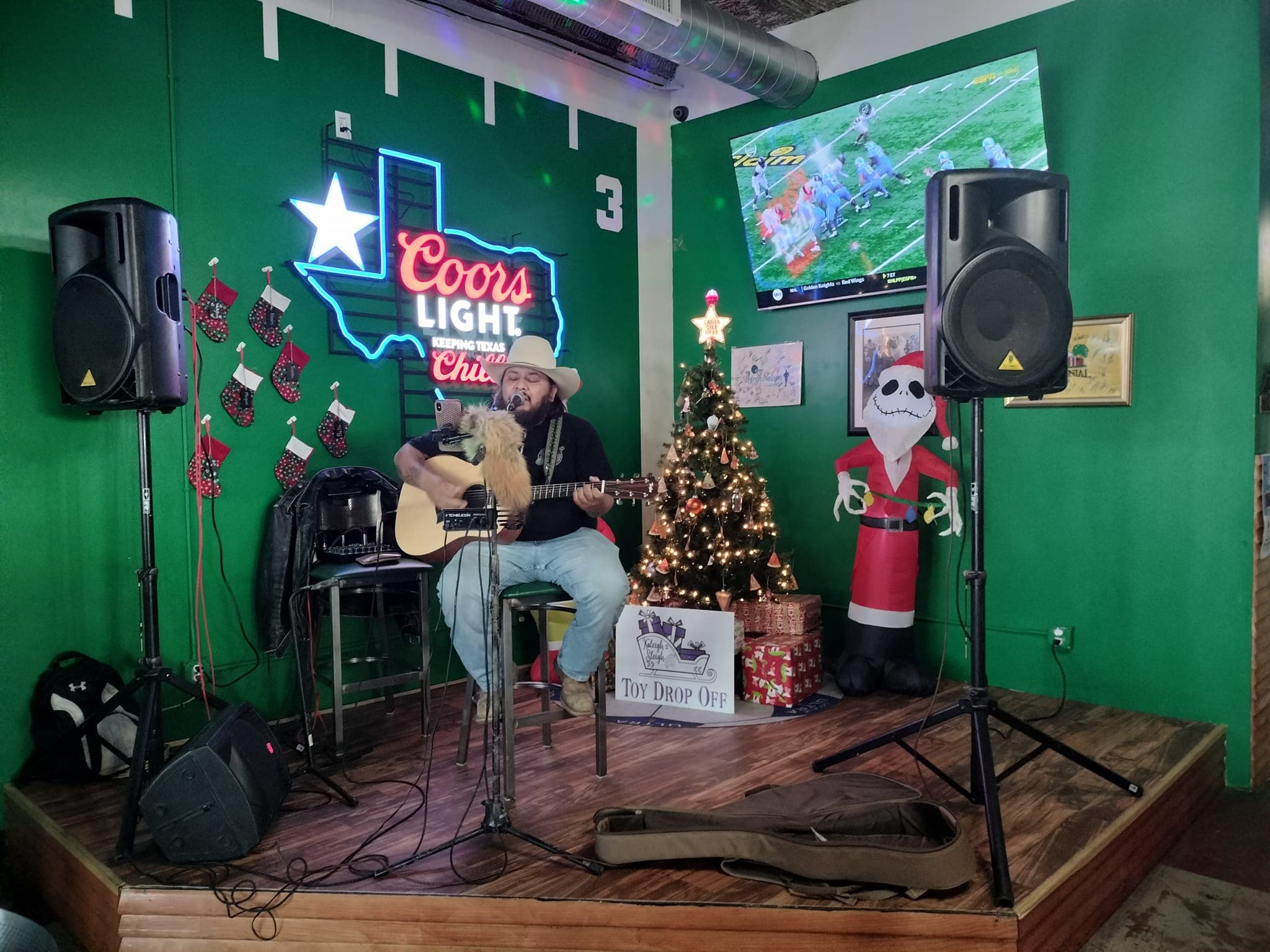 Man in cowboy hat plays guitar on a stage with Christmas decorations and neon signs.