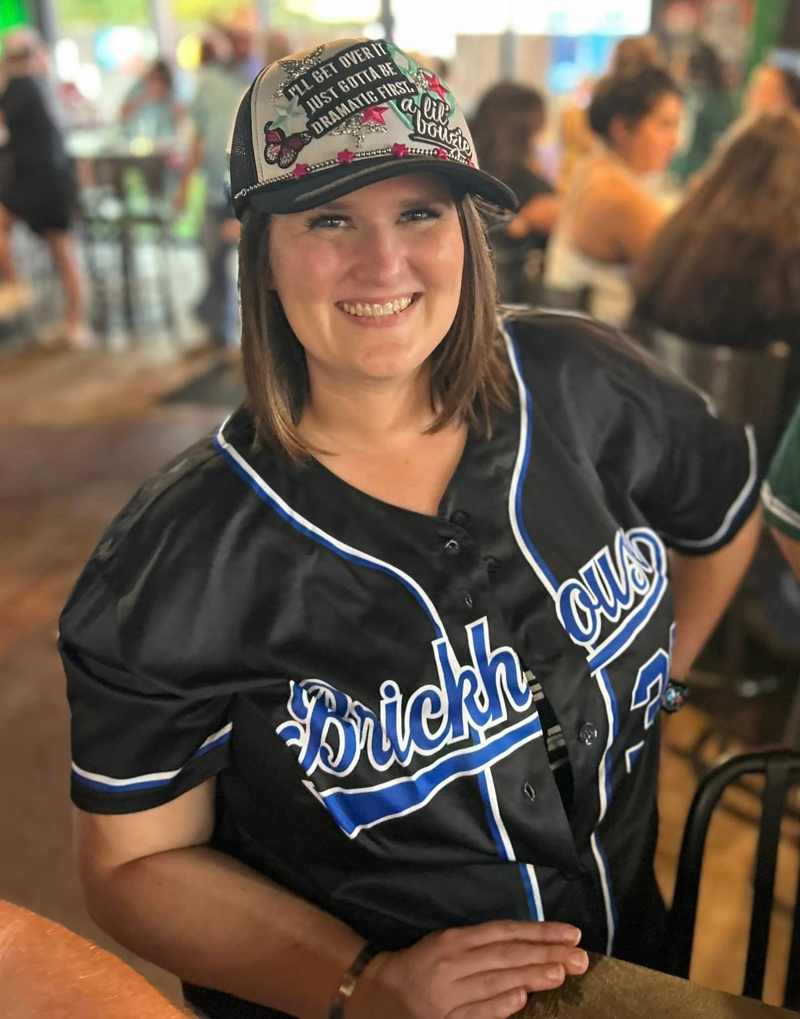 Smiling woman wearing a black baseball jersey and a decorated trucker hat in a restaurant.