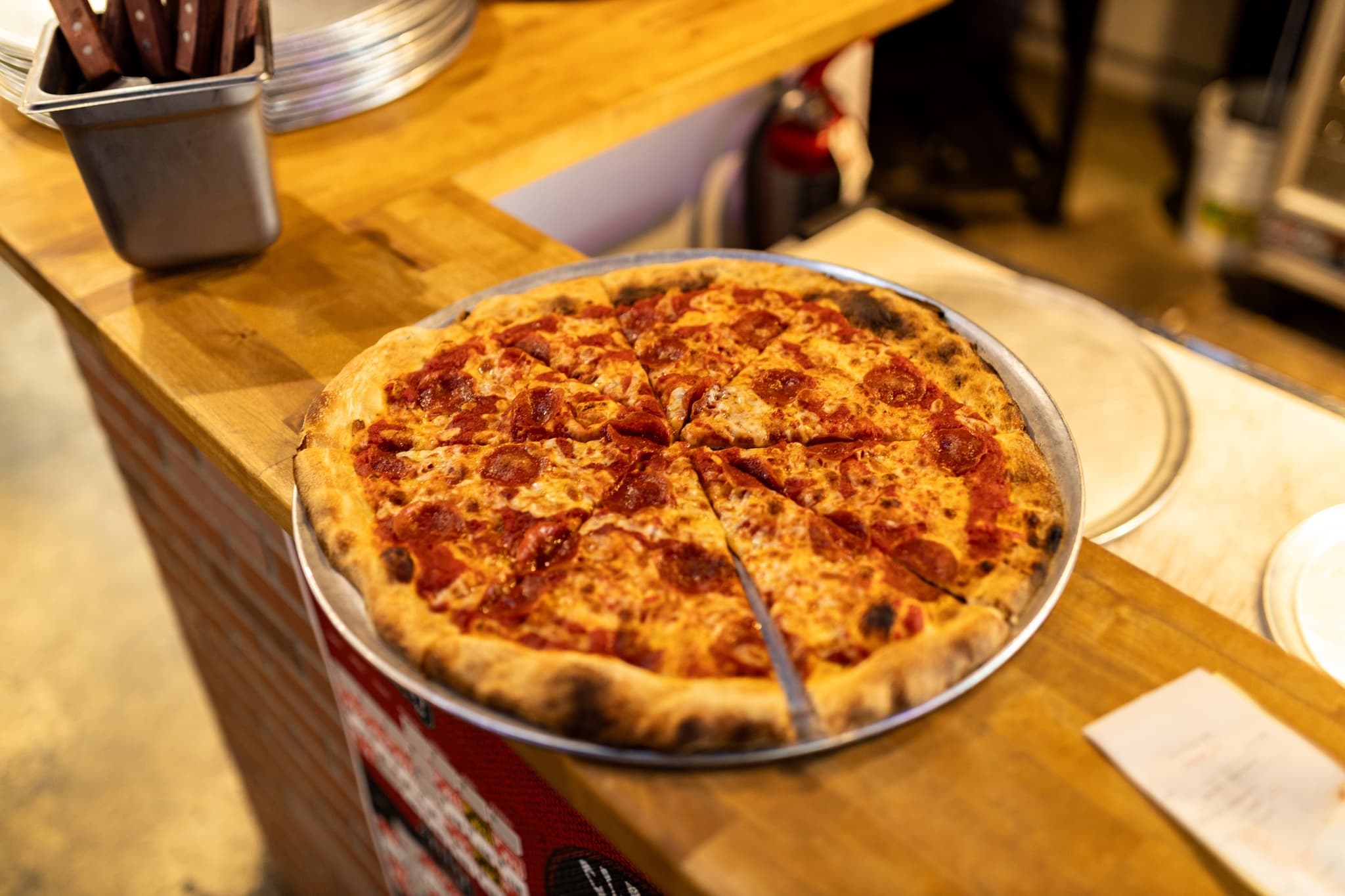 Sliced pepperoni pizza with a charred crust on a metal tray atop a wooden counter.