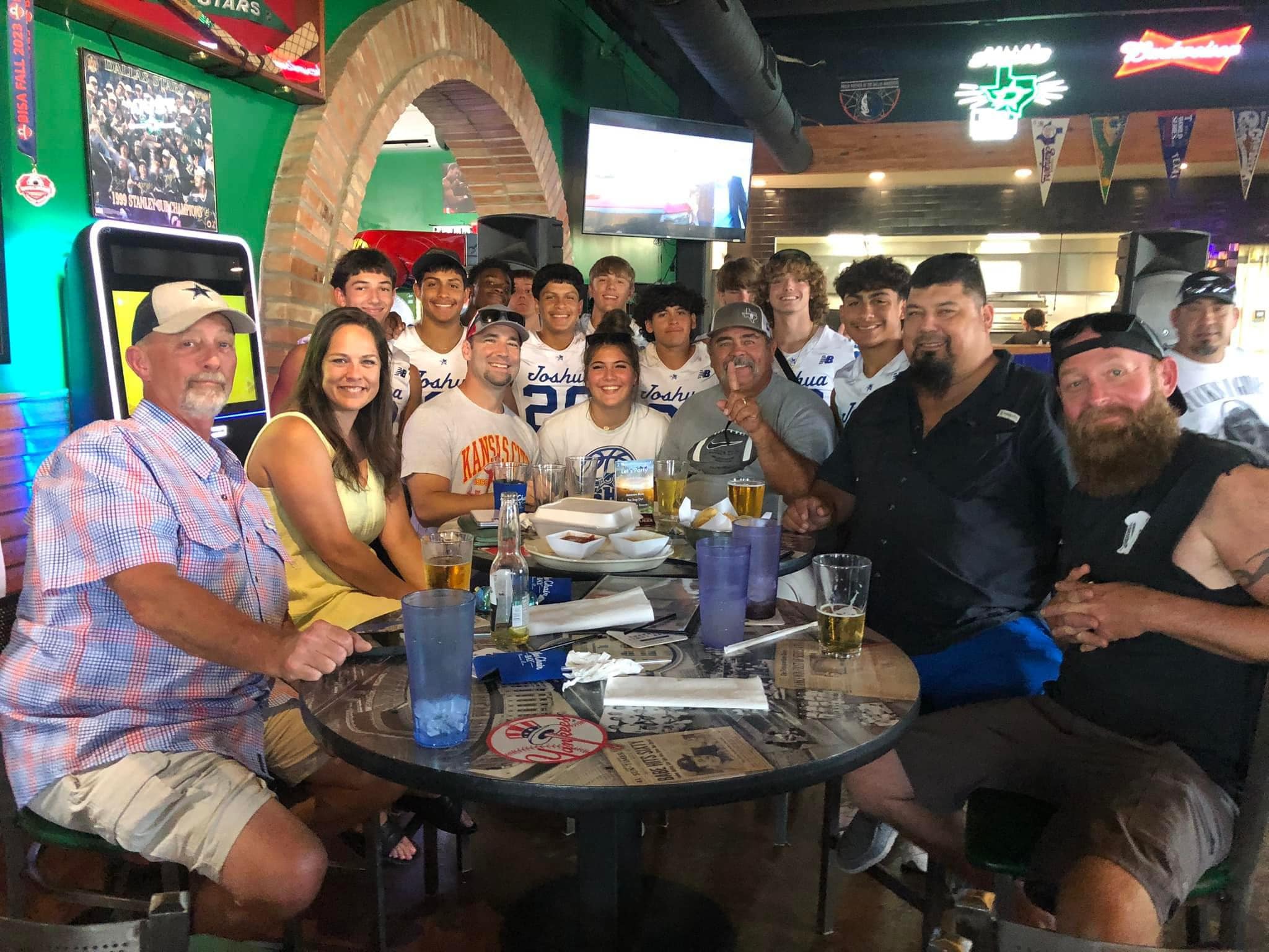 Adults and youth athletes in jerseys pose around a table in a sports bar.