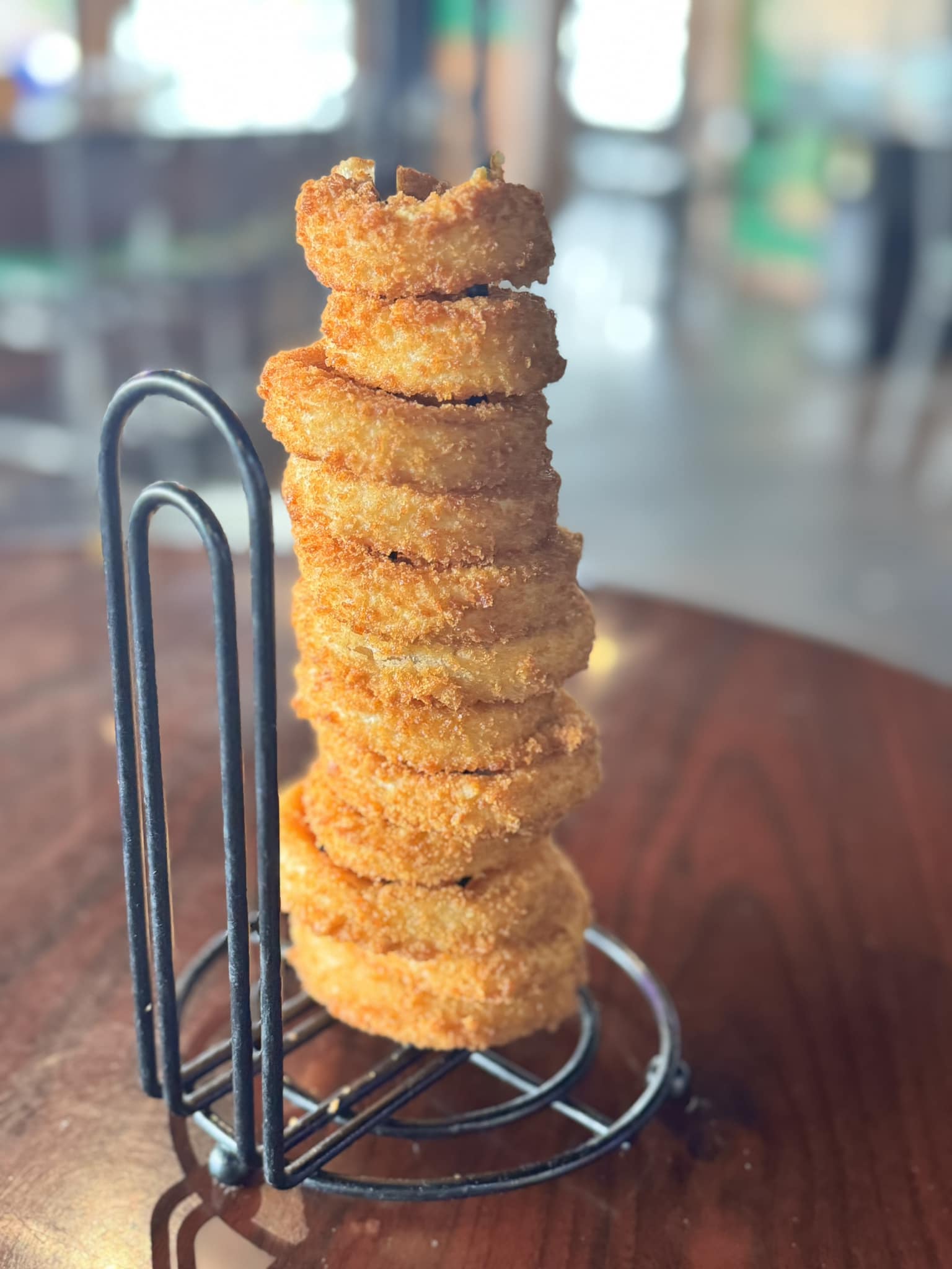 A tall stack of crispy, golden-brown breaded onion rings on a black wire stand.