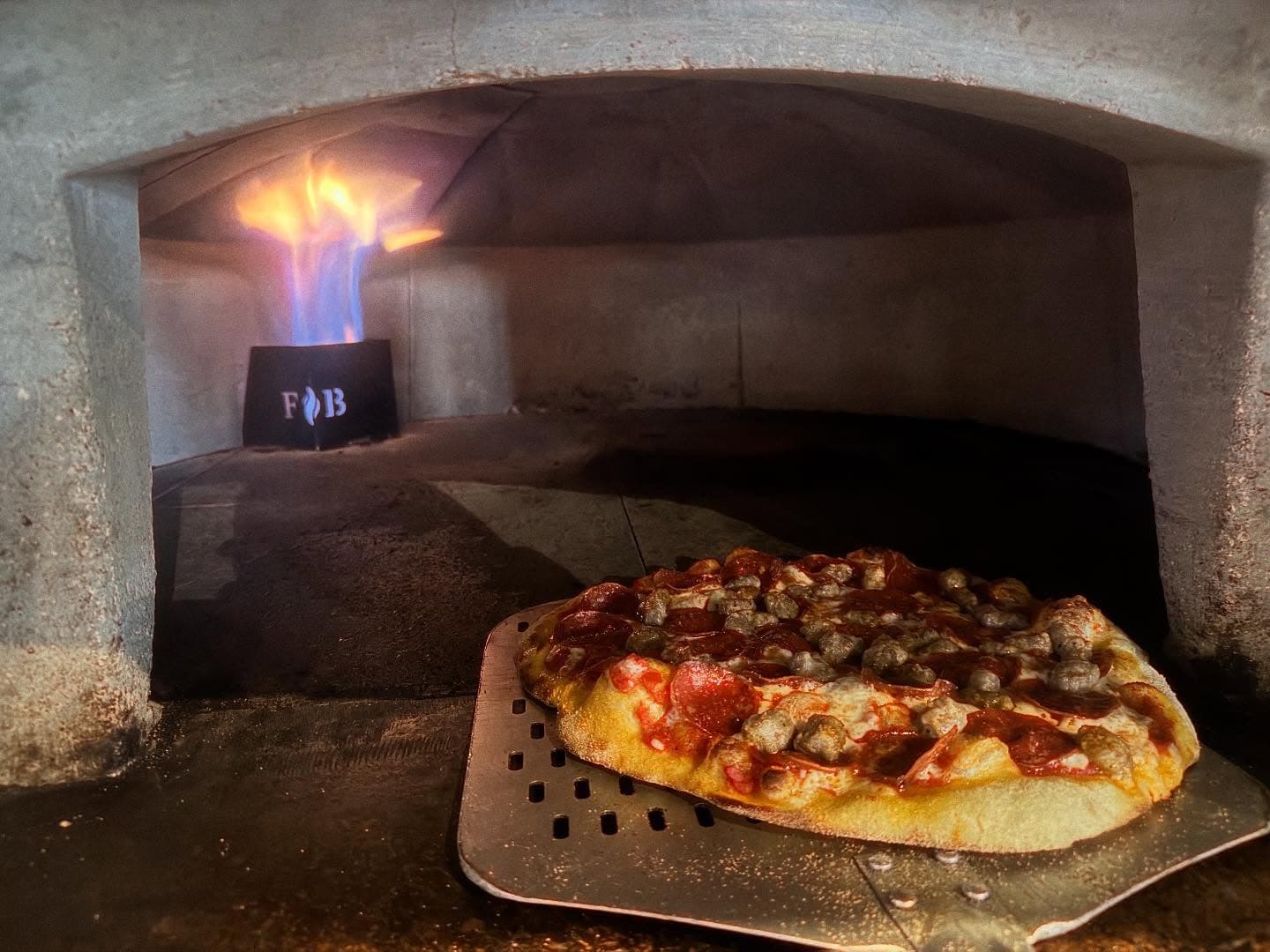 Pepperoni and sausage pizza on a metal peel inside a stone oven with flames.