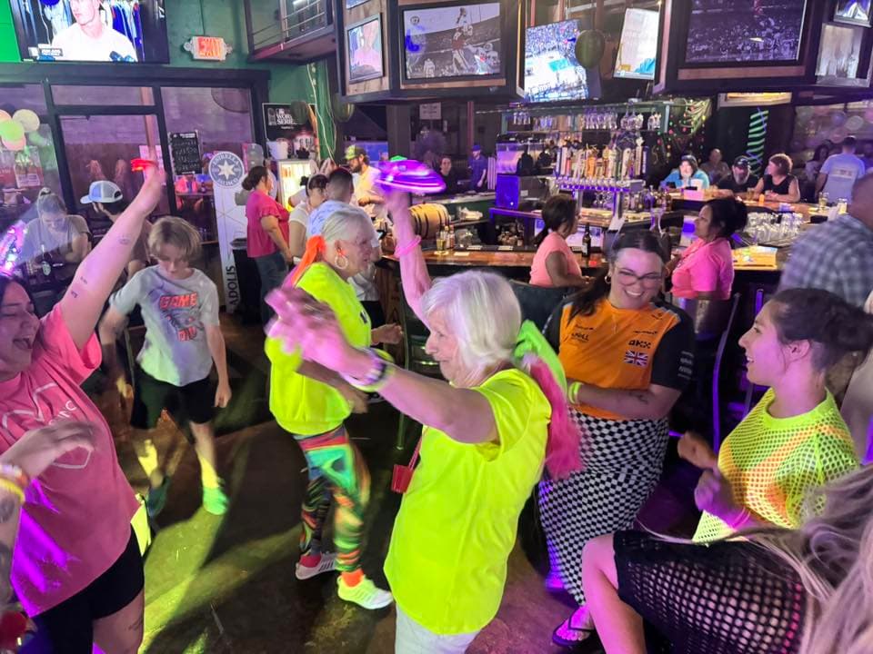 Diverse group of people in neon clothing dancing and celebrating in a vibrant sports bar.