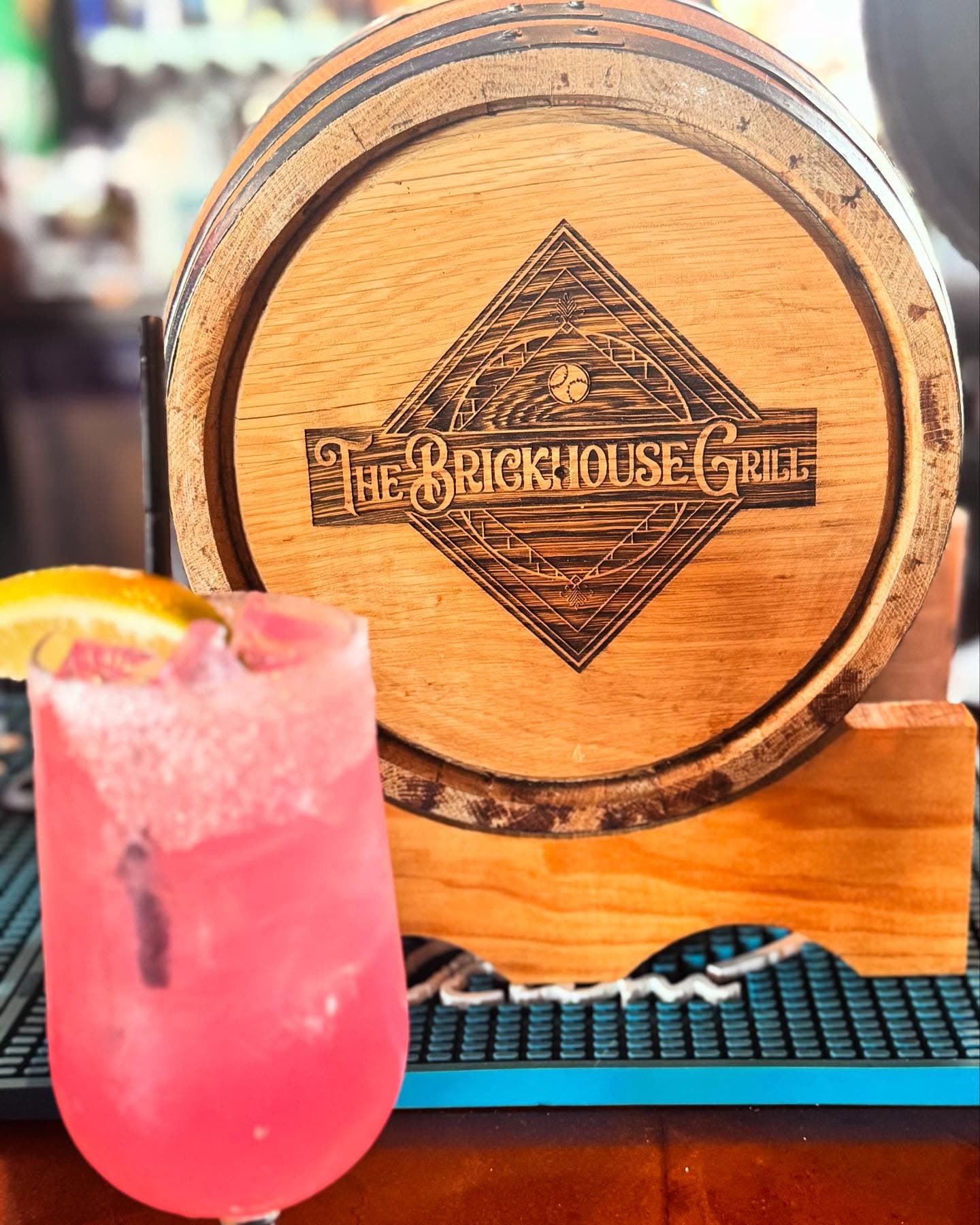 Pink cocktail with citrus garnish before a wooden barrel head featuring The Brickhouse Grill logo.