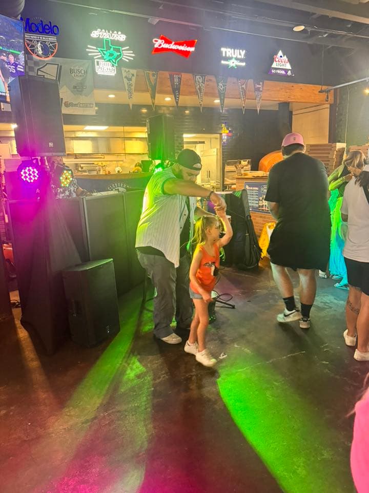 Man and young girl dance in a bar featuring neon signs and colorful DJ lights.