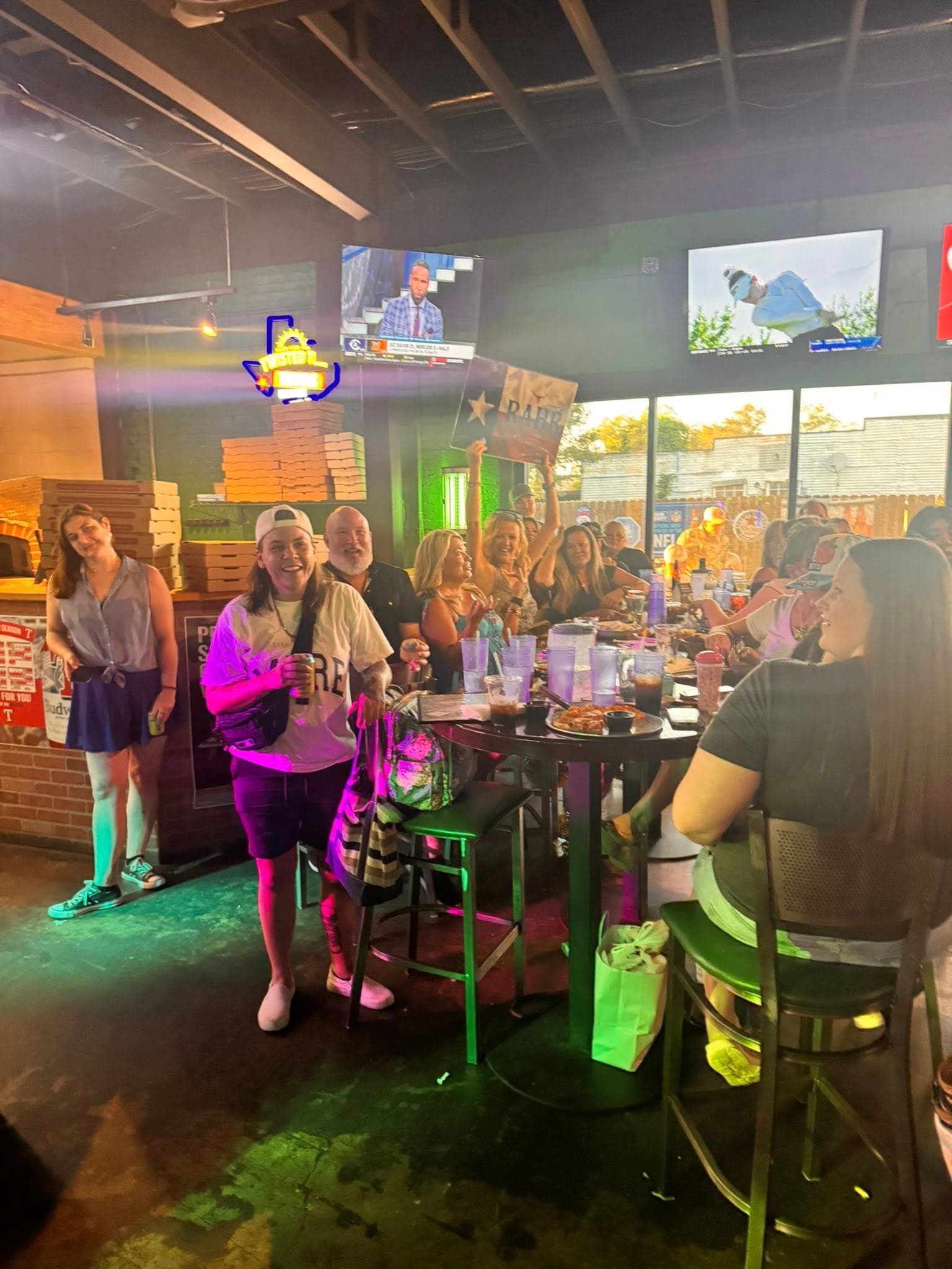 People cheering at a bar table with pizza and drinks under colorful neon lights.