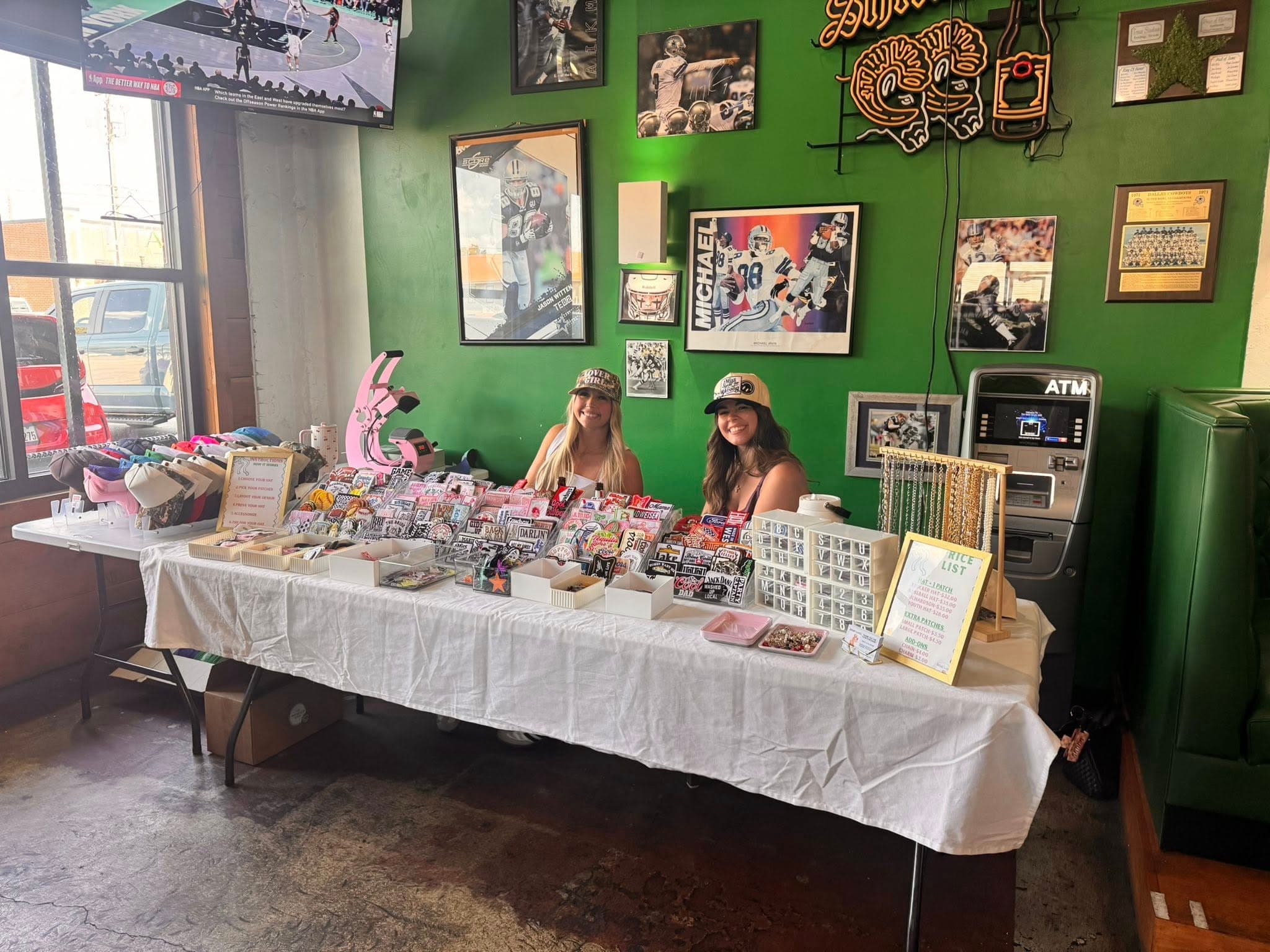 Two women smile behind a table displaying custom hats and patches in a sports bar.