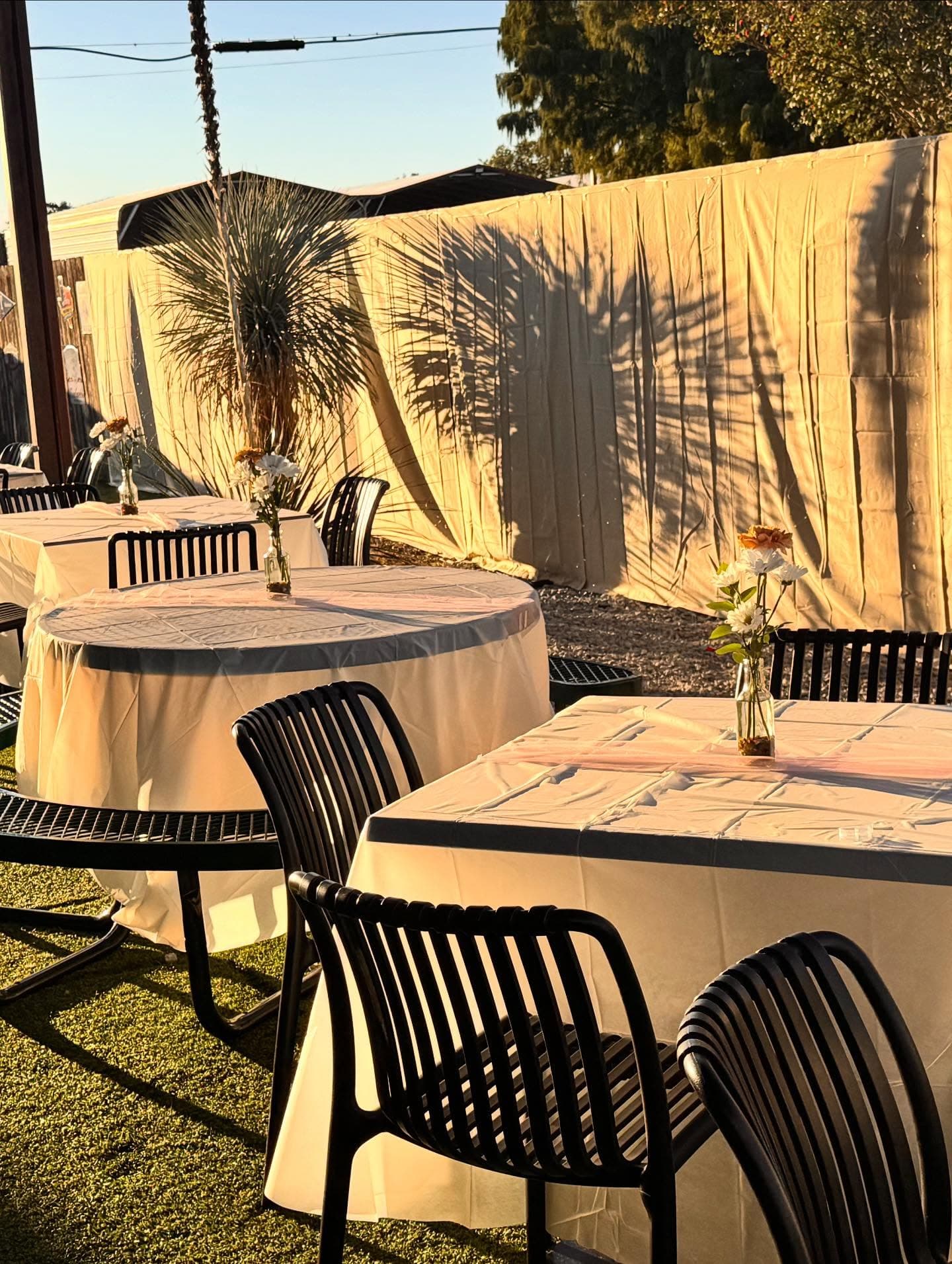 Outdoor event tables with white tablecloths and floral centerpieces under warm, golden hour sunlight.