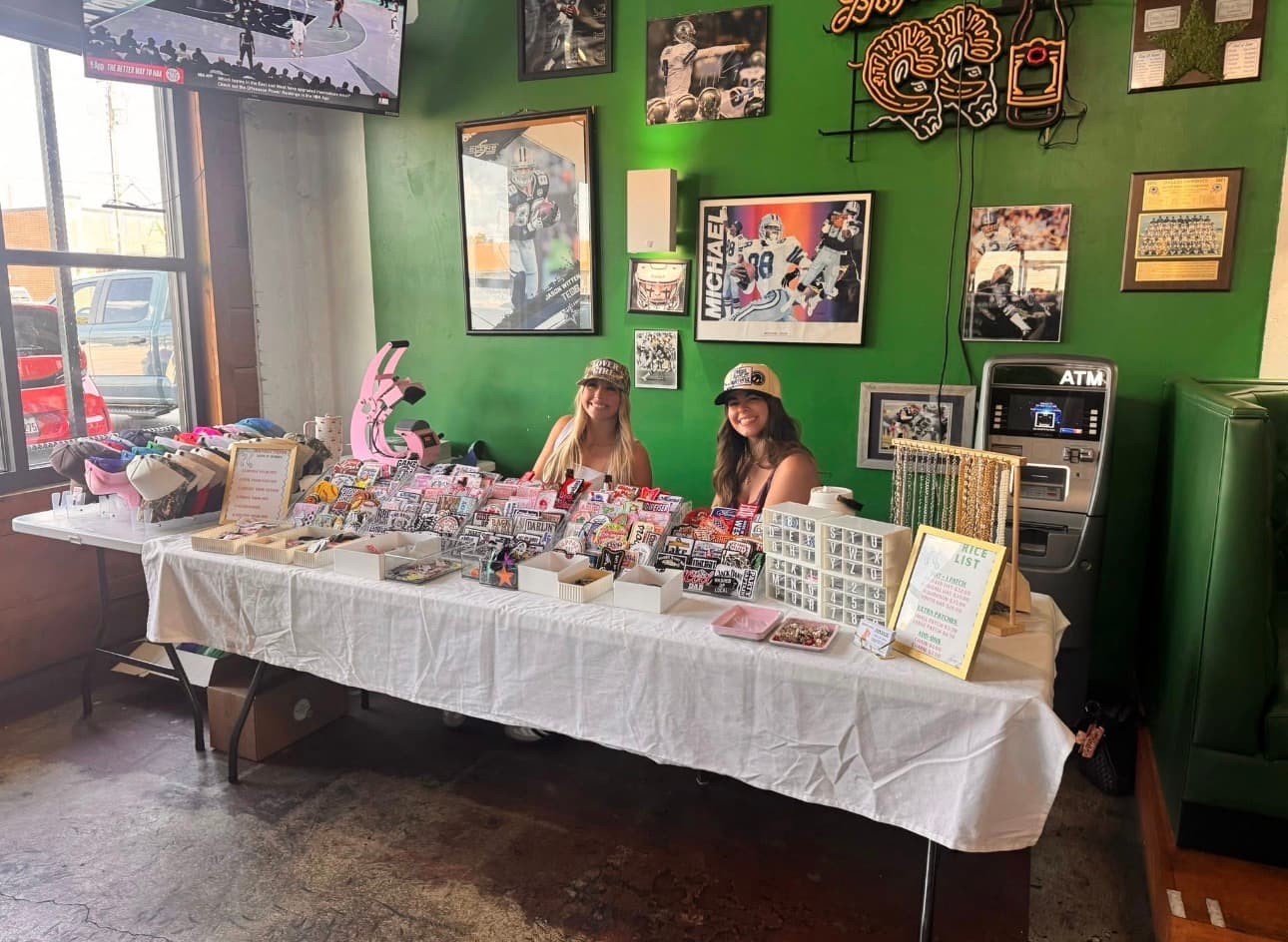 Two women smiling behind a table of patches and jewelry in a sports bar.