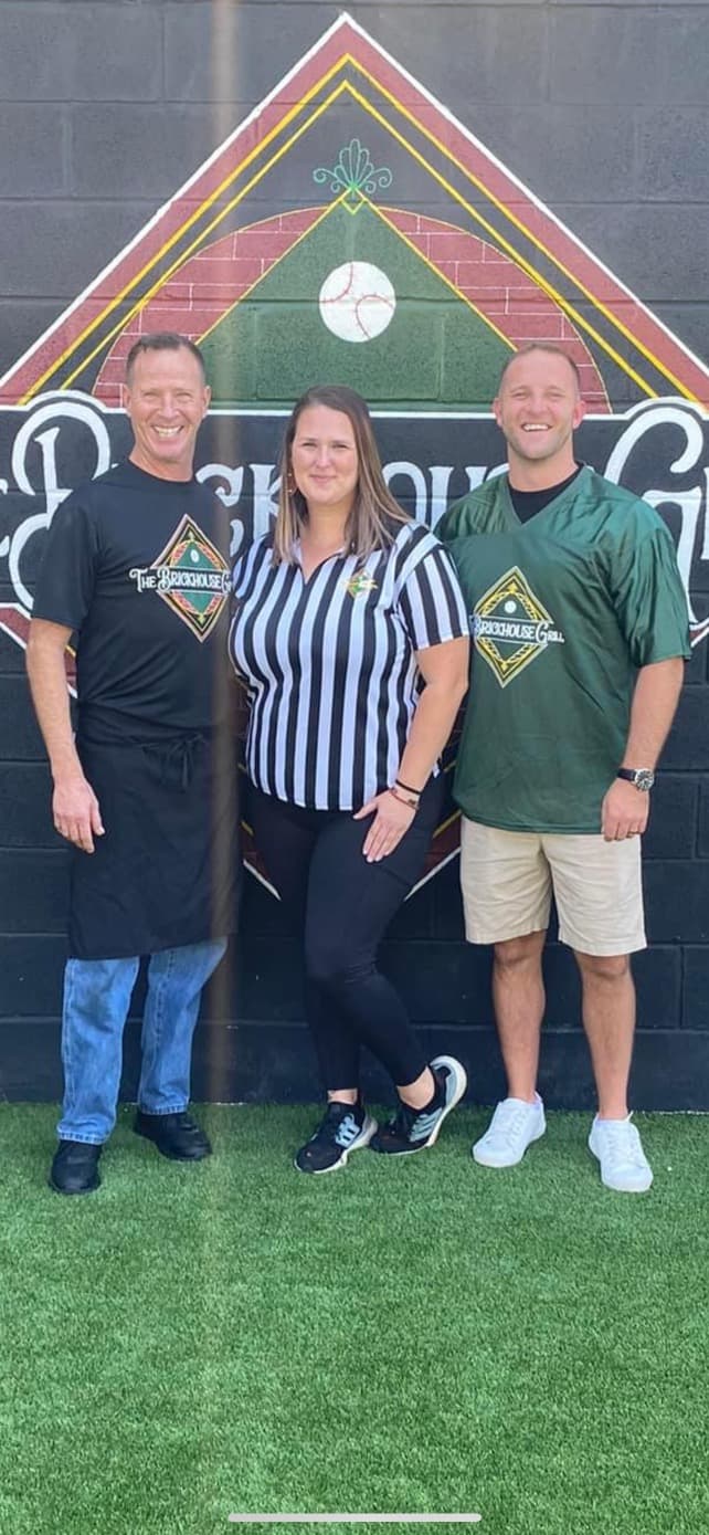 Three smiling people in sports-themed clothing stand before a large Brickhouse Grill logo.