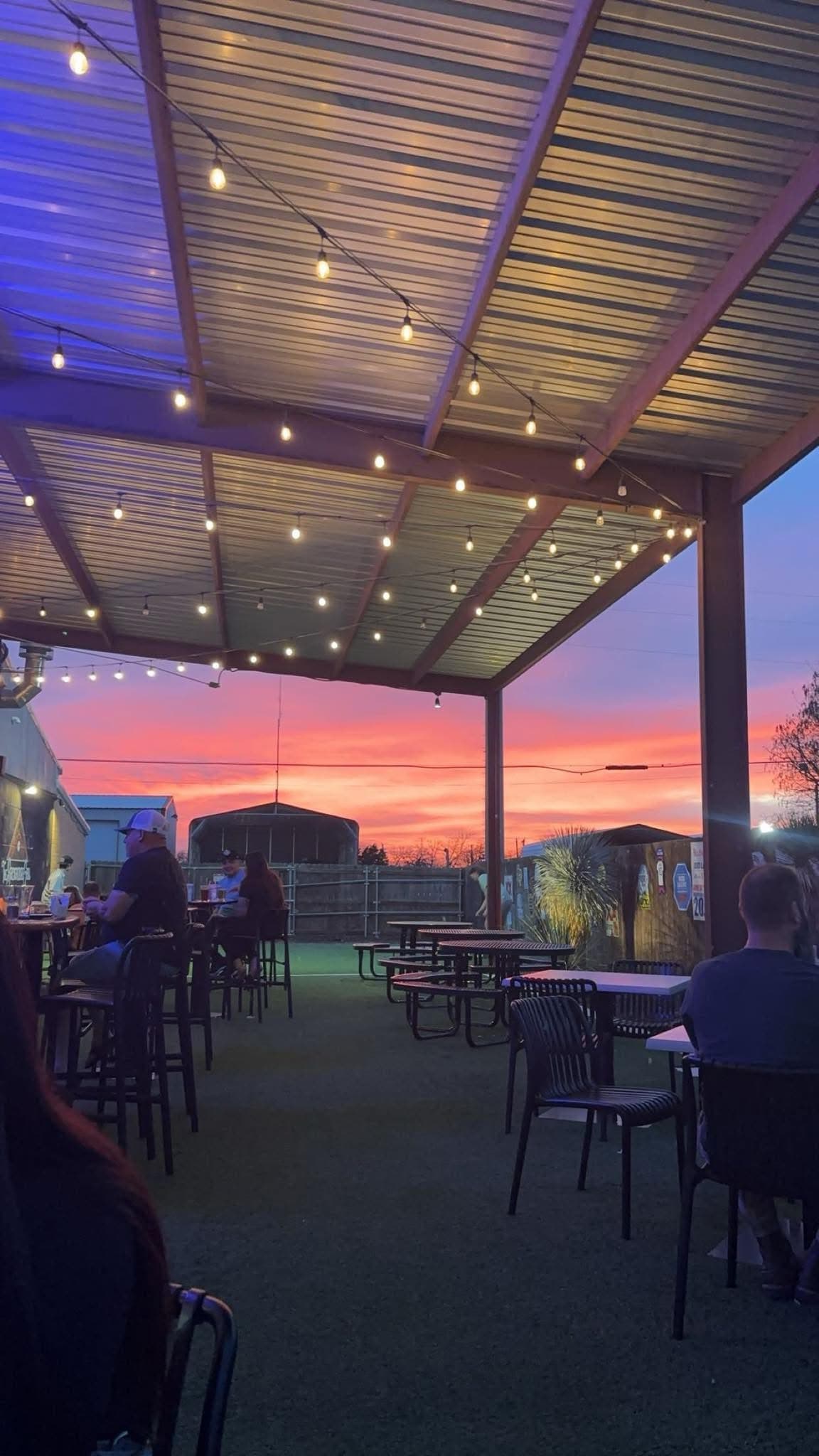 Outdoor patio with string lights and people seated under a vibrant pink and orange sunset.