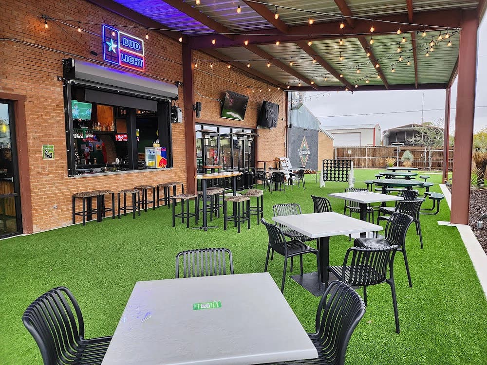 Outdoor patio with artificial turf, white tables, black chairs, and a neon Bud Light sign.