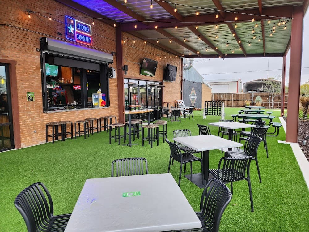 Outdoor bar patio with artificial turf, white tables, black chairs, and overhead string lights.